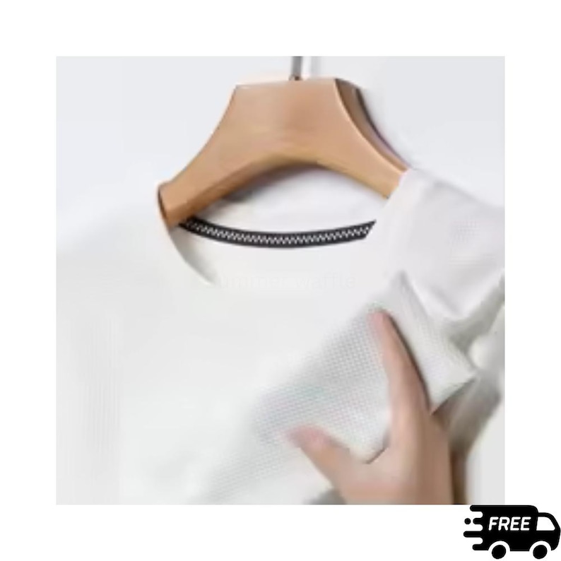 May include: A white, short-sleeved shirt with a textured pattern, hanging on a wooden hanger. The shirt has a black trim around the neckline. A hand is touching the shirt.