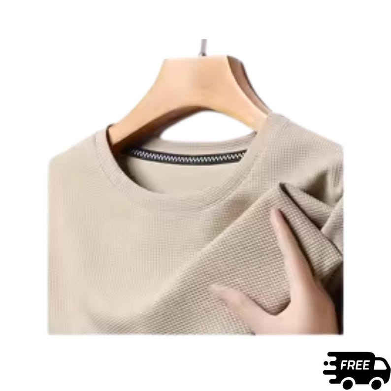 May include: A beige waffle-knit crew neck shirt hanging on a wooden hanger. The shirt has a textured surface and a black and white trim around the neckline. The sleeve is being held by a hand.