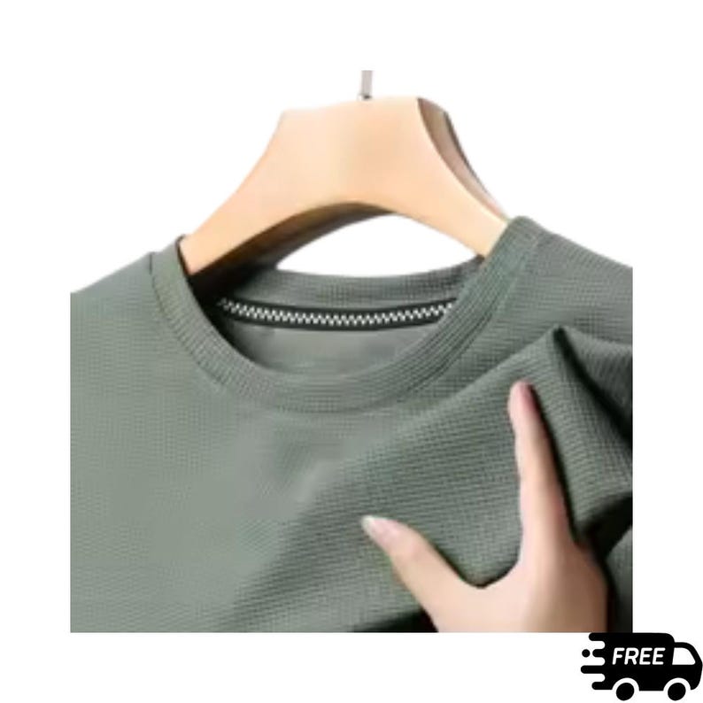 May include: A sage green crew neck shirt with a textured waffle knit pattern. The shirt is displayed on a wooden hanger. The neckline features a black and white zipper detail. A hand is holding the shirt.