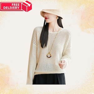 Può includere: Maglione color crema a maglia aperta, con maniche lunghe e scollo tondo. Il maglione è abbinato a pantaloni neri. È visibile una lunga collana dorata con un ciondolo circolare. L'immagine include una grafica "CONSEGNA GRATUITA".