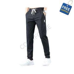 May include: Dark denim-colored pants with a drawstring waist and white drawstring. The pants have pockets and a small white tag on the side. The model is wearing white sneakers and a silver watch. The pants are a casual style.