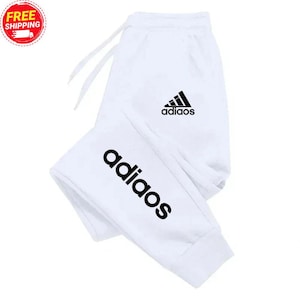 Puede incluir: Pantalones deportivos blancos con cintura con cordón y logotipo negro. La palabra "adiaos" está impresa en letras grandes y en negrita en la pierna izquierda. El logotipo de la marca y la palabra "adiaos" también están impresos en la pierna derecha.