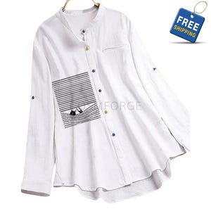 May include: White long-sleeve shirt with a mandarin collar and a pocket featuring a black and white striped design. The shirt has colorful buttons and a relaxed fit. The sleeves can be rolled up and secured with a button.