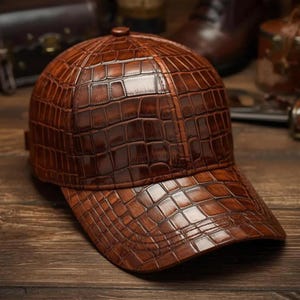 Puede incluir: Una gorra de béisbol marrón con una textura en relieve de piel de cocodrilo en toda la superficie. La gorra tiene una corona estructurada, una visera curva y un botón en la parte superior central, con un acabado brillante en tonos marrón oscuro.