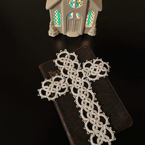 May include: A white, intricately designed cross rests on a closed, brown book. The cross is made of a delicate, woven material. In the background, a small, white church model with colorful stained-glass windows is visible.
