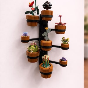 Op de afbeelding: Een zwarte wandplantenstandaard met verschillende kleine potplanten. De planten omvatten rode bloemen, vetplanten en cactussen, allemaal in bruine potten. De standaard is gemaakt van zwart metaal en is aan een witte muur bevestigd.