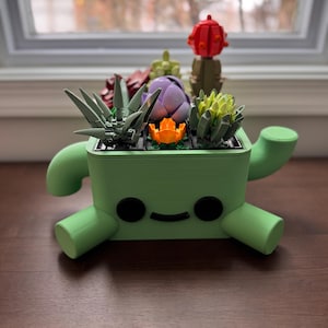 May include: A light green, rectangular planter shaped like a character with two black circular eyes and a curved mouth. The planter holds a collection of colourful plastic building block succulents in shades of red, purple, orange, yellow, and green.