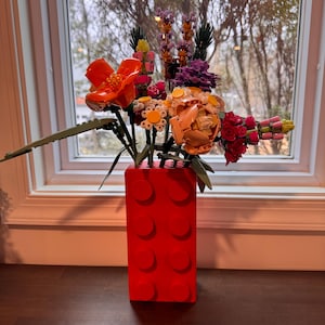 May include: A vibrant arrangement of colorful Lego flowers in a bright red Lego brick vase. The flowers include orange, pink, yellow, and purple blooms, with green stems and leaves. The vase is rectangular with circular studs.