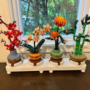 Pode incluir: Um conjunto de quatro plantas de blocos de plástico para montar em vasos individuais, incluindo um ramo com flores vermelhas, uma orquídea laranja-claro, um crisântemo laranja-brilhante e um caule de bambu verde sobre uma base branca.