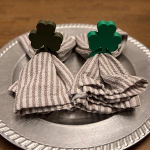 May include: Two cloth napkins with brown and white stripes are held by shamrock-shaped napkin rings. One ring is dark green, the other is bright green. The napkins are on a silver plate.