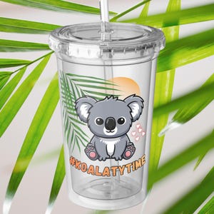 May include: Clear plastic tumbler with a lid and straw. The tumbler features a cartoon koala bear illustration with the text "#KOALATYTIME". The koala is grey with pink accents, set against a backdrop of a sunset and palm leaves.