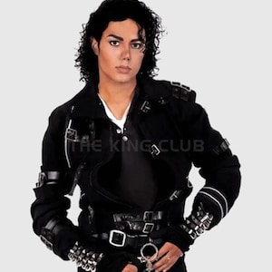 Puede incluir: Chaqueta negra con múltiples hebillas, correas y detalles plateados. La chaqueta tiene un cuello alto y se usa sobre una camisa blanca. El texto "THE KING CLUB" es visible en la chaqueta.