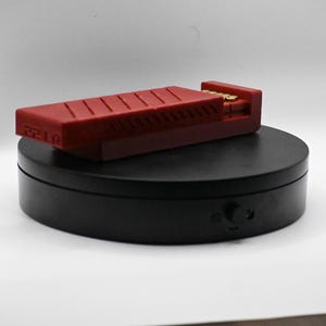 May include: A red rectangular object with a ridged top, possibly a tool or accessory, rests on a black circular base. The object has the number "2210" printed on its side. The base has a small control knob.