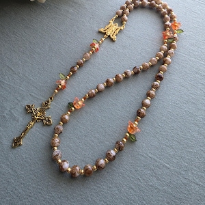 May include: A rosary featuring brown and white marbled beads interspersed with small gold-tone spacers and translucent orange flower-shaped beads with green leaves. The piece includes a gold-tone metal centerpiece and a matching ornate crucifix.