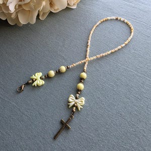 May include: A beaded lanyard featuring a mix of cream and beige seed beads, pale yellow round beads, and two pale yellow bow charms. The accessory includes a bronze-toned metal cross pendant at one end and a matching lobster claw clasp.