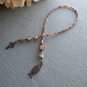 May include: A beaded loop bracelet featuring small orange, brown, and grey glass seed beads. The bracelet includes larger, textured amber-colored glass beads and two bronze-toned metal charms: one shaped like a cross and one shaped like a fish with the word JESUS embossed on it.