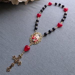 May include: A rosary featuring black round beads and translucent red heart-shaped beads on a gold-tone chain. The centerpiece is an oval pendant depicting the Sacred Hearts of Jesus and Mary in a filigree frame, ending in a decorative gold cross.