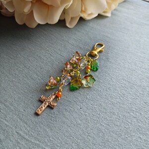 May include: A gold-colored keychain charm featuring a cross pendant and a cluster of colorful beads and charms. The charm includes orange, green, and clear beads, along with leaf-shaped and teardrop-shaped charms. The clasp is also gold-colored.