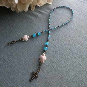 May include: A beaded bookmark featuring small turquoise and brown seed beads, translucent light blue glass beads, and two carved white turtle charms. The bookmark is finished with a bronze-toned metal anchor charm at one end and a hook at the other.
