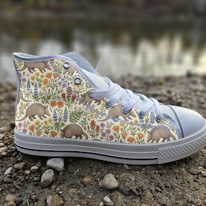 Puede incluir: Una zapatilla de caña alta con una parte superior de lona color crema con un patrón repetitivo de armadillos marrones, amapolas naranjas, lupinos morados y helechos verdes. El calzado tiene cordones gris claro, puntera de goma gris y suela gris.