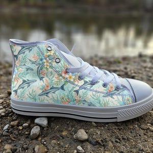 May include: A high-top trainer featuring a light blue background with a repeating pattern of grey sharks, orange coral, and white flowers. The shoe has a white rubber toe cap, a white rubber sole with a thin black stripe, and white laces.