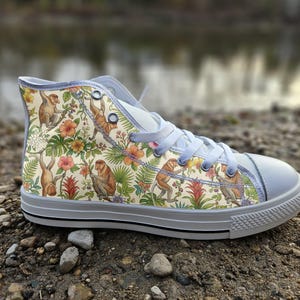 May include: A high-top trainer featuring a cream-coloured canvas upper with a repeating pattern of brown monkeys, green tropical leaves, and vibrant orange and pink hibiscus flowers. The shoe has light grey laces, a matching toe cap, and a rubber sole.