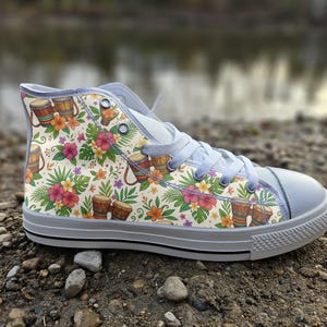 Peut inclure: Une basket montante avec une tige en toile crème ornée d'un motif répétitif de djembés marron, de fleurs d'hibiscus roses et jaunes éclatantes, et de feuilles de palmier tropicales vertes. La chaussure possède des lacets gris clair et une semelle en caoutchouc blanc.