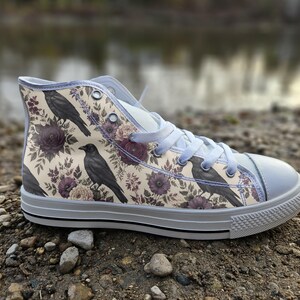 Peut inclure: Une basket montante avec une tige en toile couleur crème ornée d'un motif répétitif de corbeaux noirs et d'illustrations florales violet foncé. La chaussure comprend des lacets gris clair, un bout en caoutchouc gris et une semelle blanche.