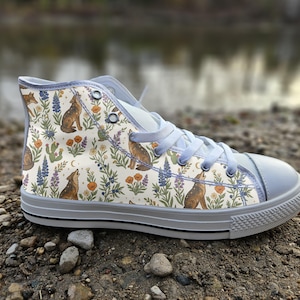 Puede incluir: Una zapatilla de caña alta con una parte superior de lona color crema con un patrón repetitivo de coyotes aullando, flores de lupino moradas, amapolas naranjas y cactus verdes. El calzado tiene cordones azul claro, puntera azul claro y suela blanca.