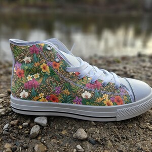 Könnte beinhalten: Ein High-Top-Sneaker mit einem lebendigen tropischen Druck aus grünem Blattwerk, gelben und roten Hibiskusblüten, violetten Orchideen und braunen Schlangen. Der Schuh hat weiße Schnürsenkel, eine weiße Gummikappe und eine weiße Sohle.