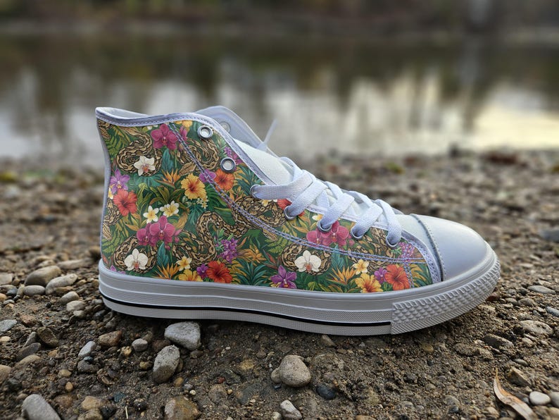 Puede incluir: Una zapatilla de ca&ntilde;a alta con un estampado tropical vibrante de follaje verde, flores de hibisco amarillas y rojas, orqu&iacute;deas moradas y serpientes marrones. El calzado tiene cordones blancos, puntera de goma blanca y suela blanca.