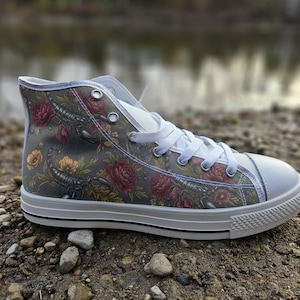 Puede incluir: Una zapatilla de caña alta con parte superior de lona gris y un estampado detallado de escorpiones negros, peonías rojas y flores amarillas. El calzado incluye cordones blancos, ojales metálicos plateados, puntera de goma blanca y suela blanca.
