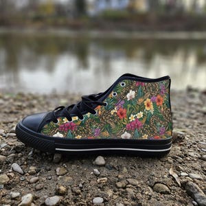 Puede incluir: Una zapatilla de ca&ntilde;a alta con puntera de goma negra, cordones negros y suela negra con una raya blanca. La parte superior de lona muestra un vibrante estampado tropical con follaje verde, texturas de piel de serpiente y flores.