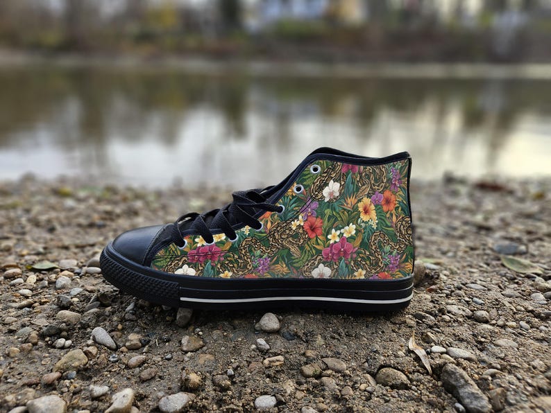 Puede incluir: Una zapatilla de ca&ntilde;a alta con puntera de goma negra, cordones negros y suela negra con una raya blanca. La parte superior de lona muestra un vibrante estampado tropical con follaje verde, texturas de piel de serpiente y flores.