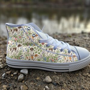 May include: A high-top sneaker featuring a cream-colored canvas upper with a repeating pattern of brown and white hunting dogs interspersed with colorful wildflowers, green foliage, light blue laces, and a solid light gray rubber toe cap and sole.