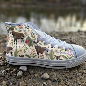 Può includere: Una sneaker alta con tomaia in tela color crema caratterizzata da un motivo ripetuto di okapi, fiori rosa e foglie verdi. La scarpa presenta lacci azzurri, occhielli in metallo argentato, punta in gomma grigio chiaro e suola grigia.