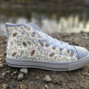 Puede incluir: Una zapatilla de caña alta con una parte superior de lona color crema que presenta un patrón repetitivo de cucarachas marrones, pequeñas flores azules y hojas verdes. El calzado incluye cordones azul claro, ojales de metal plateado, puntera y suela de goma blanca.