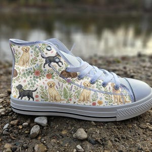 May include: A high-top trainer featuring a cream-coloured canvas upper with a repeating pattern of yellow and black Labrador dogs interspersed with colourful floral illustrations, light blue laces, and a white rubber toe cap and sole.