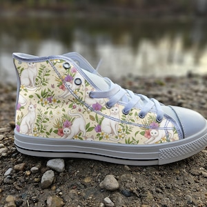 May include: A high-top sneaker with a cream-colored canvas upper featuring a repeating pattern of white cats, green leaves, and small pink and yellow flowers. The shoe includes light gray laces, a matching gray rubber toe cap, and a gray sole.