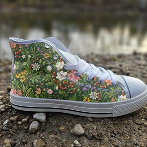May include: A high-top trainer featuring a dark green canvas upper with a detailed botanical print of various flowers, green leaves, and small snakes. The shoe includes white laces, a white rubber toe cap, and a white sole with a thin black stripe.