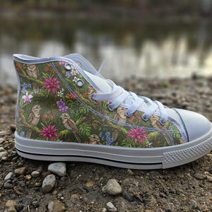 May include: A high-top sneaker featuring a green background with a repeating pattern of brown tawny frogmouth birds, vibrant pink flowers, and green tropical leaves. The shoe has white laces, a white rubber toe cap, and a white sole with black trim.
