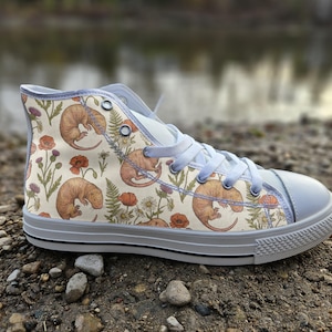May include: A high-top sneaker with a cream-colored canvas upper featuring a repeating pattern of small brown mole-like creatures, orange poppies, and green ferns. The shoe has white laces, a white rubber toe cap, and a white sole with black trim.