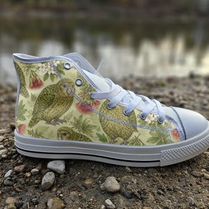 May include: A high-top trainer featuring a light yellow canvas upper with a repeating pattern of green owls, white flowers, and pink botanical elements. The shoe includes white laces, silver metal eyelets, a white rubber toe cap, and a white sole.