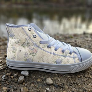 May include: A high-top trainer featuring a cream-coloured canvas upper with a repeating pattern of white dogs, blue hydrangea flowers, and green foliage. The shoe includes light blue laces, silver metal eyelets, and a white rubber toe cap and sole.