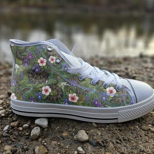 Könnte beinhalten: Ein High-Top-Sneaker mit grünem botanischem Muster, braunen Schlangen, violetten Schwertlilien und rosa Hibiskusblüten. Der Schuh hat weiße Schnürsenkel, eine weiße Gummikappe und eine weiße Gummisohle mit einem dünnen schwarzen Streifen.