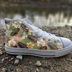 Könnte beinhalten: Ein High-Top-Sneaker mit hellgrünem Hintergrund und einem wiederkehrenden Muster aus braunen Kakerlaken, gelben Orchideen und grünen Palmblättern. Der Schuh hat weiße Schnürsenkel, eine weiße Gummikappe und eine weiße Sohle mit schwarzem Streifen.