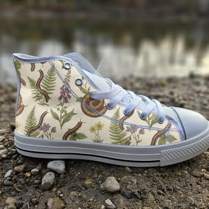 May include: A high-top trainer with a cream-coloured canvas upper featuring a botanical and insect print. The pattern includes brown millipedes, green fern fronds, purple flowers, and a brown ammonite fossil. It has light grey laces and a rubber sole.