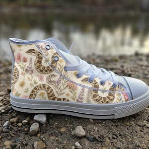 May include: A high-top sneaker featuring a cream-colored canvas upper with a repeating pattern of brown snakes and various wildflowers in shades of pink, yellow, and purple. The shoe includes light gray laces, a matching toe cap, and a rubber sole.