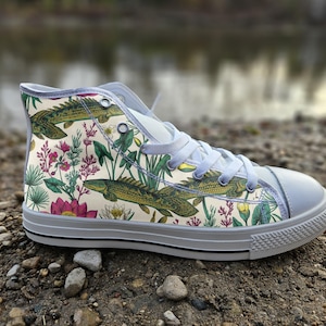 May include: A high-top sneaker featuring a cream-colored canvas upper with a botanical and aquatic print. The design includes green fish, pink lotus flowers, and various water plants. It has a white rubber toe cap, white laces, and a white sole.