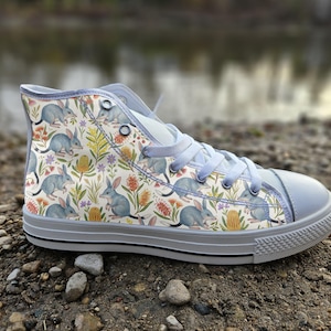 May include: A high-top sneaker featuring a repeating pattern of grey bilbies, yellow banksia flowers, and orange bottlebrush plants on a white background. The shoe has light grey laces, a matching grey rubber toe cap, and a grey sole with a black stripe.